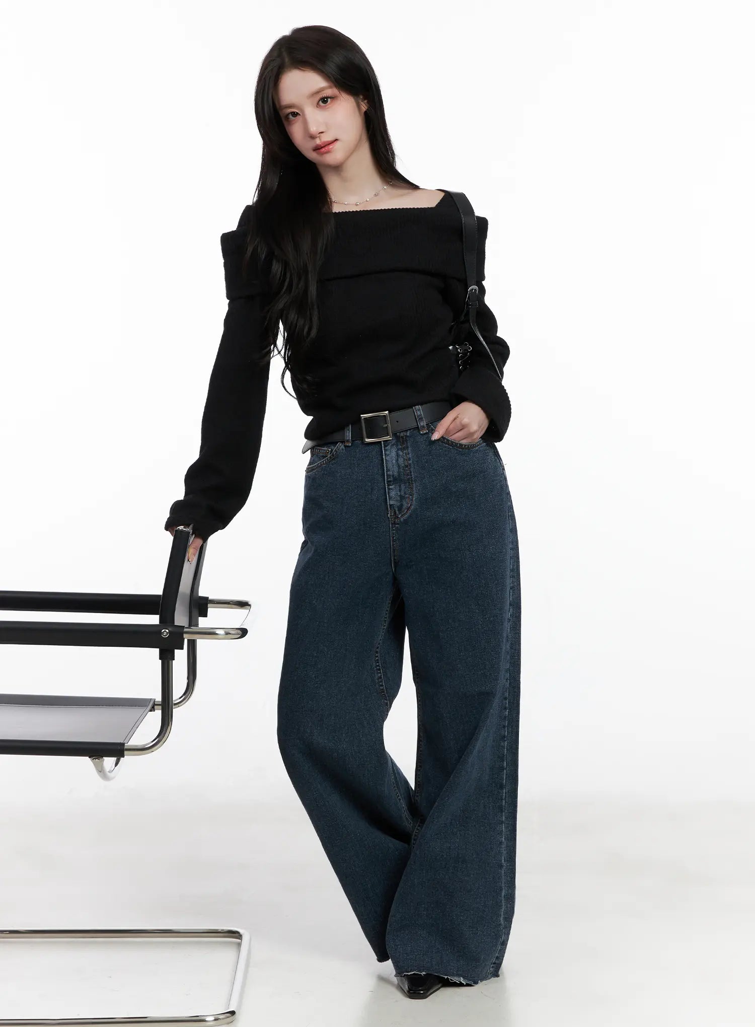 Louisa Ultra Wide Flared Jeans CN527 in dark blue, full-body editorial shot on model beside a steel chair, wide flared denim with black belt, paired with black off-shoulder knit and black crossbody bag.