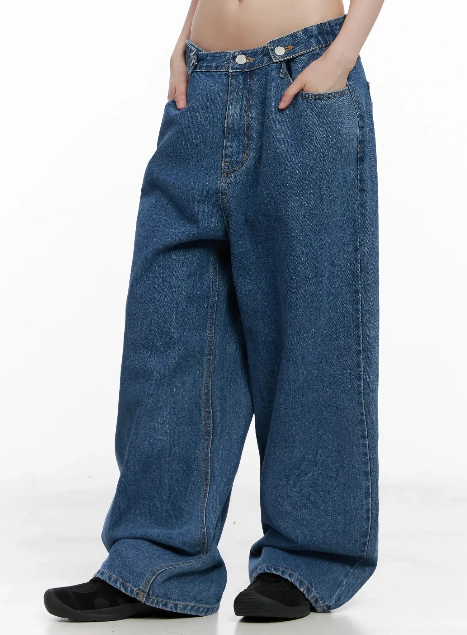Pharita Loose-Fit Jeans IL528 in mid blue, side view on model, wide-leg silhouette with deep side pocket and diagonal crossover waistband visible, floor-grazing hem length.