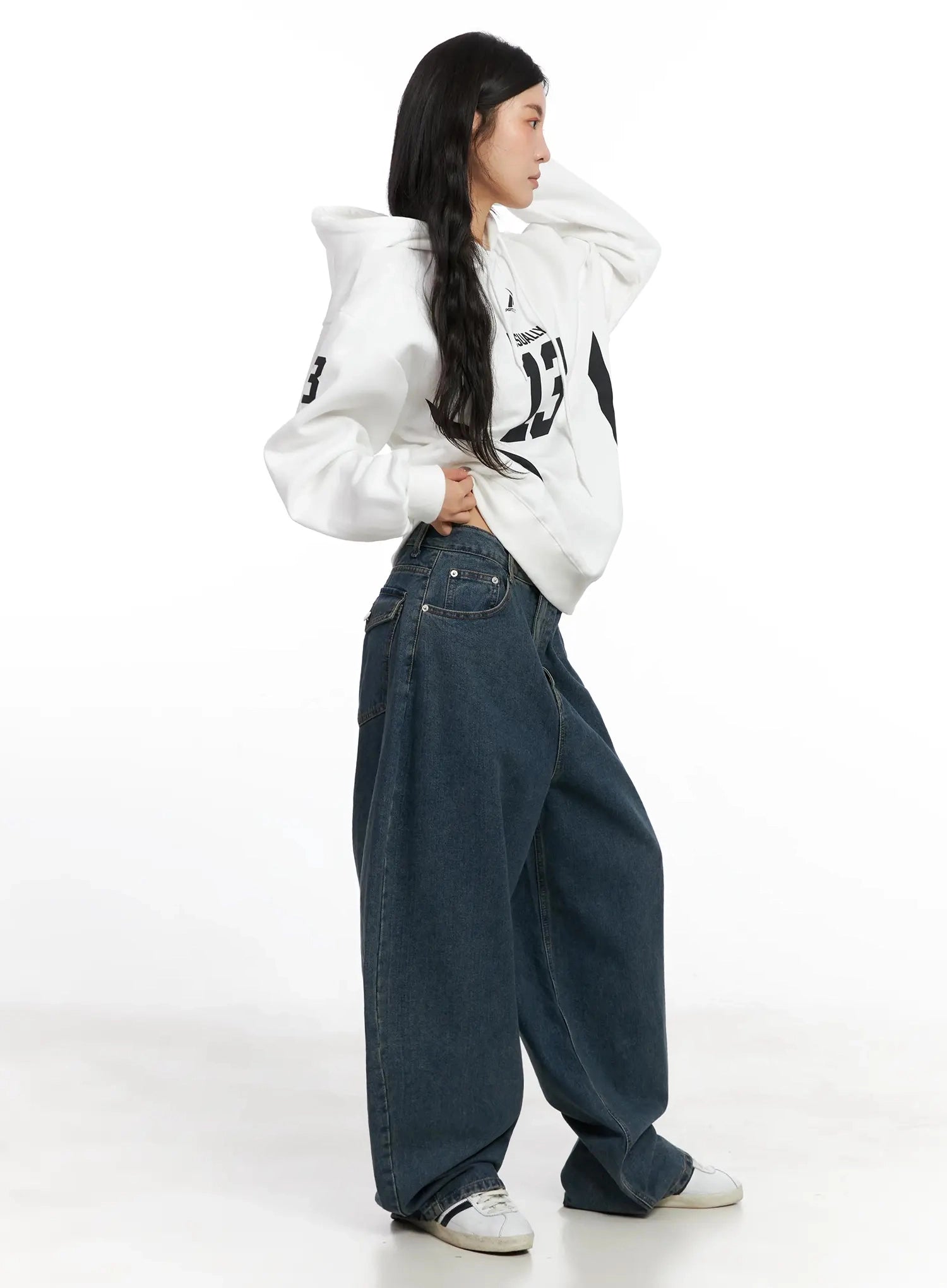Aleena Mid-Wash Baggy Jeans IN528 in dark blue, three-quarter side shot on model in white graphic hoodie, dynamic pose showing voluminous leg shape and mid-wash cotton denim.