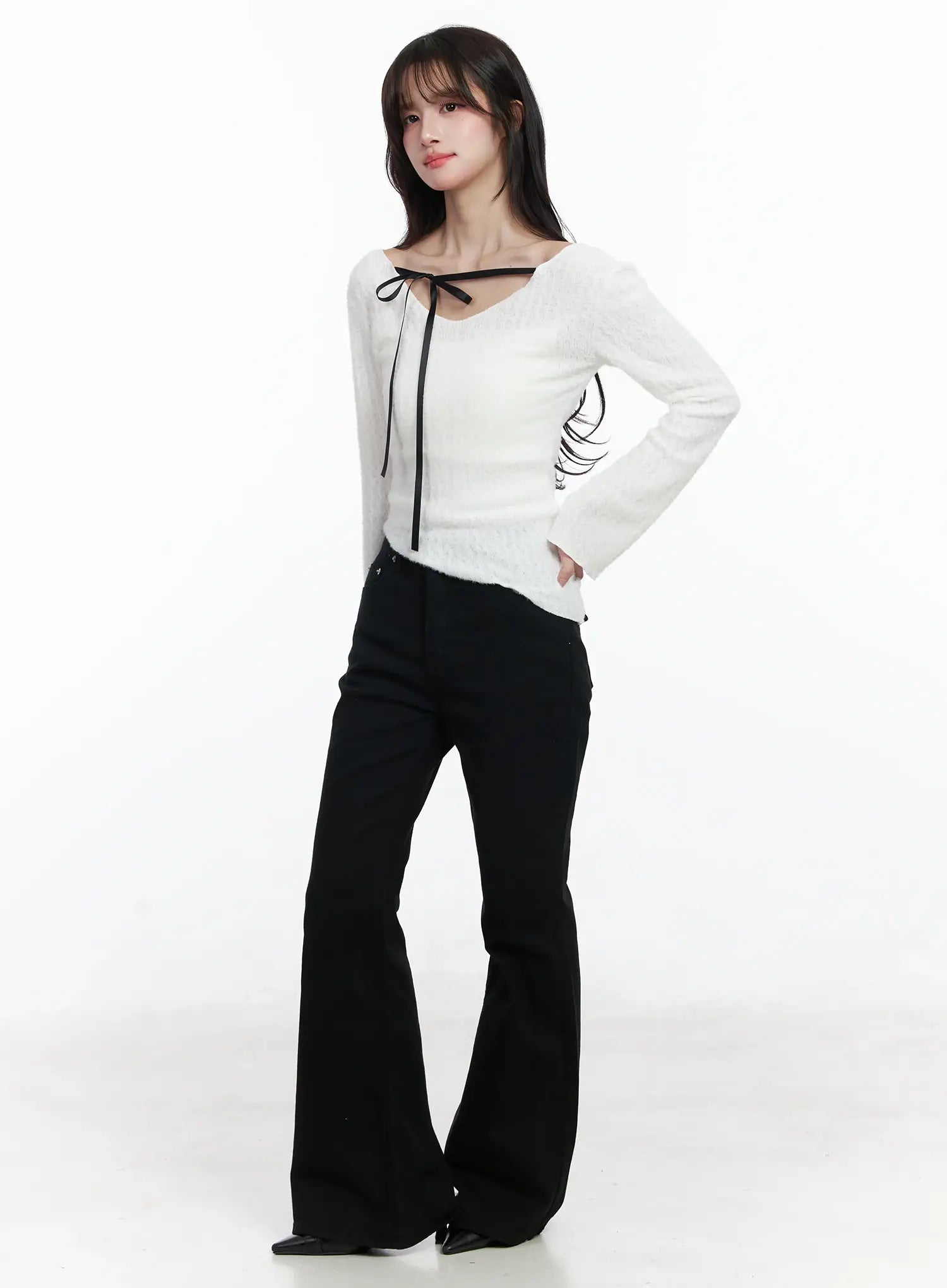 Ellis High-Waist Bootcut Jeans IJ529 in black, full-body 3/4-angle editorial shot on model, bootcut flare silhouette styled with white bow-tie fuzzy knit top and black pointed-toe heels.