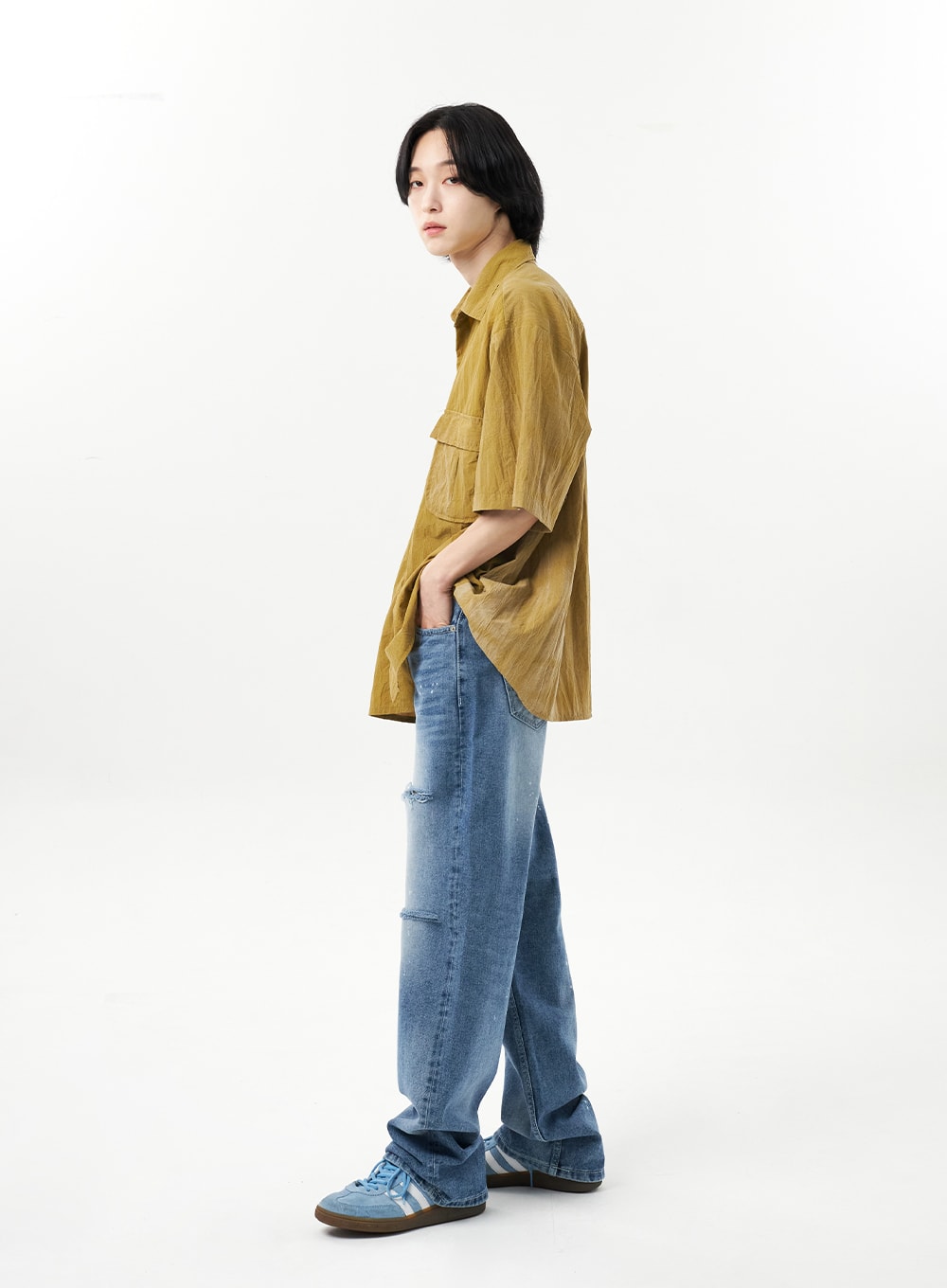Ripped Jeans Unisex CU315 in mid blue, full-body side view on male model, wide-leg denim silhouette with side-visible rips at knee, relaxed cotton fit, paired with olive oversized shirt.