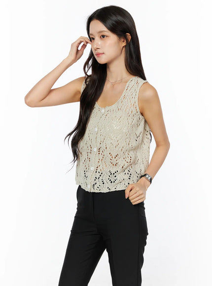 Crochet Knit Sleeveless Top CU522 in beige, side-front view on model with hand raised to face, open-knit crochet lace pattern, scoop neckline, paired with black trousers.