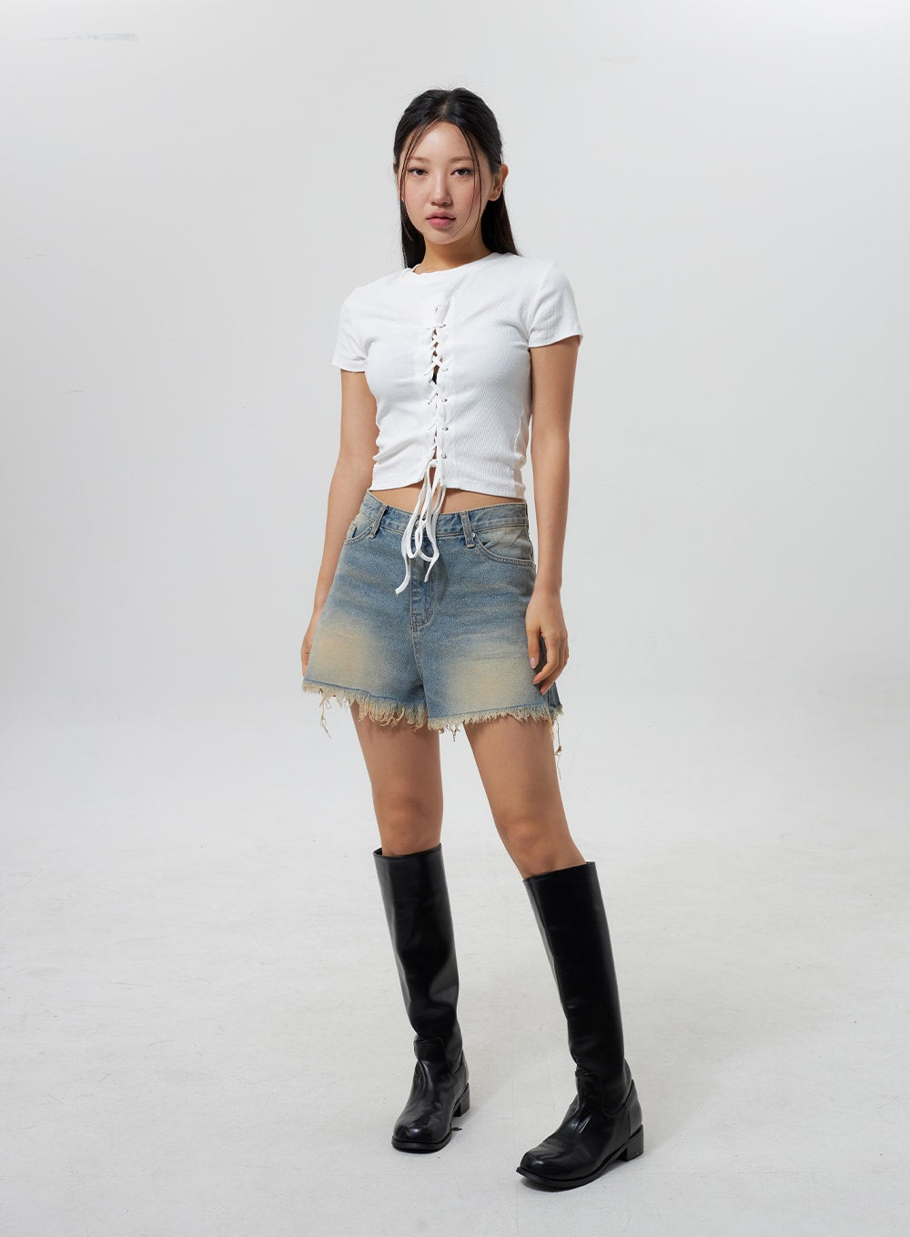 Mid-Rise Ripped Jeans CY323 in light blue, full-body slight angle on model, mid-rise ripped denim shorts with yellow-toned fade and raw hem, styled with white lace-up crop top and knee-high boots.