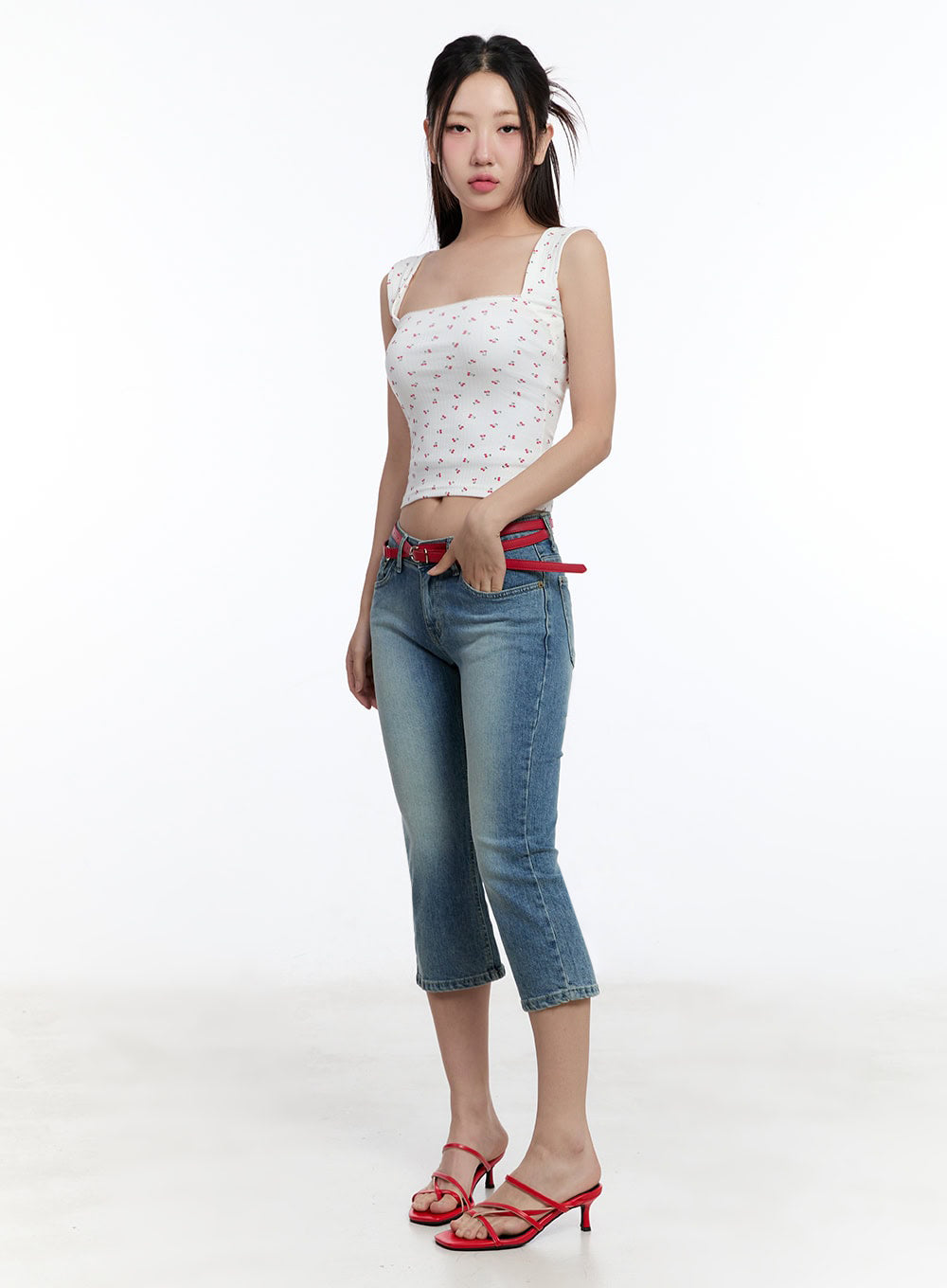 Kyra Cropped Low-Rise Skinny Jeans IU503 in blue, three-quarter front angle on model, washed denim low-rise with red belt threaded through loops, paired with white floral tank and red strappy mules.