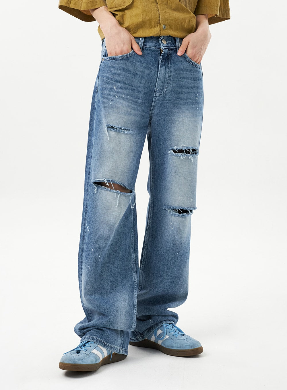 Ripped Jeans Unisex CU315 in mid blue, front thigh-to-ankle close-up on model hands in pockets, wide-fit cotton denim with multiple destroyed rips and heavy fraying, faded wash with paint splatter.
