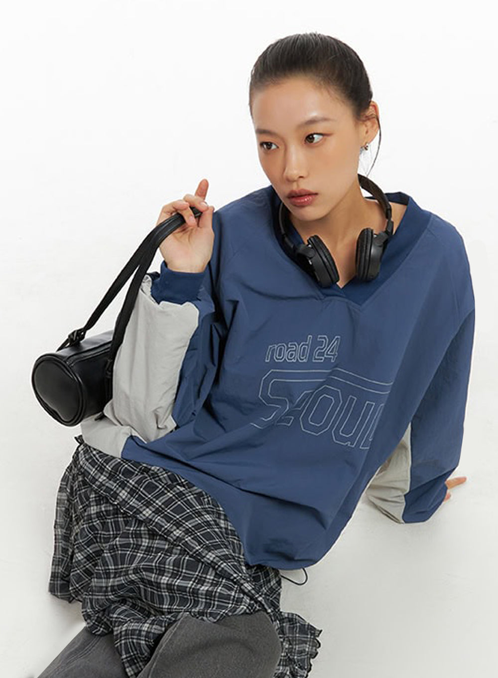 Emery Cotton Strap Baggy Jeans IO409 in dark gray, editorial seated upper-body shot on model, jeans partially visible at hip, layered with blue Seoul graphic top and plaid skirt.