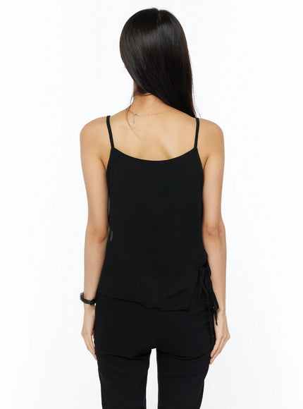 Sheer Ribbon Cami Top CU522 in black, back view on model, thin spaghetti straps, open back, side ribbon tie detail at hip, sheer layered hem, styled with black trousers.