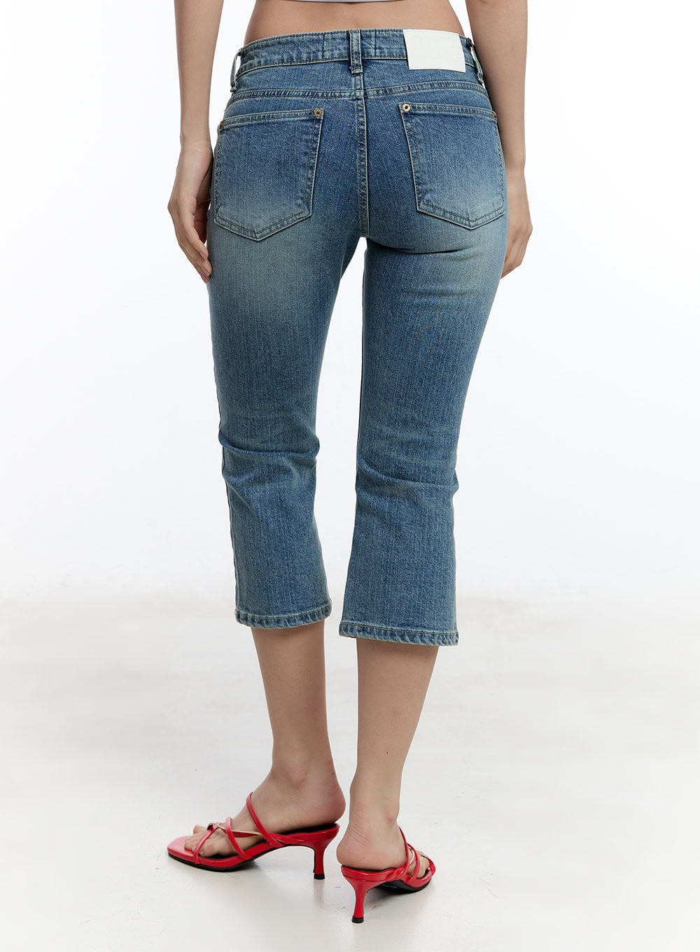 Kyra Cropped Low-Rise Skinny Jeans IU503 in blue, back view on model, washed denim showing dual back pockets with gold rivets, label at waistband, cropped hem, worn with red strappy mules.
