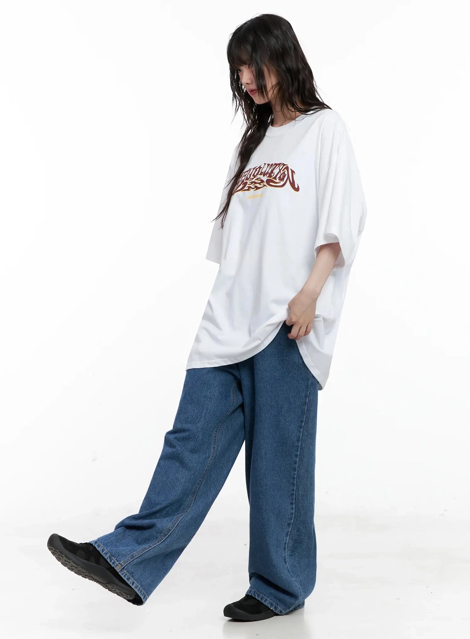 Pharita Loose-Fit Jeans IL528 in mid blue, dynamic walking side pose on model, wide-leg cotton denim with floor-length hem, styled with oversized white graphic tee.