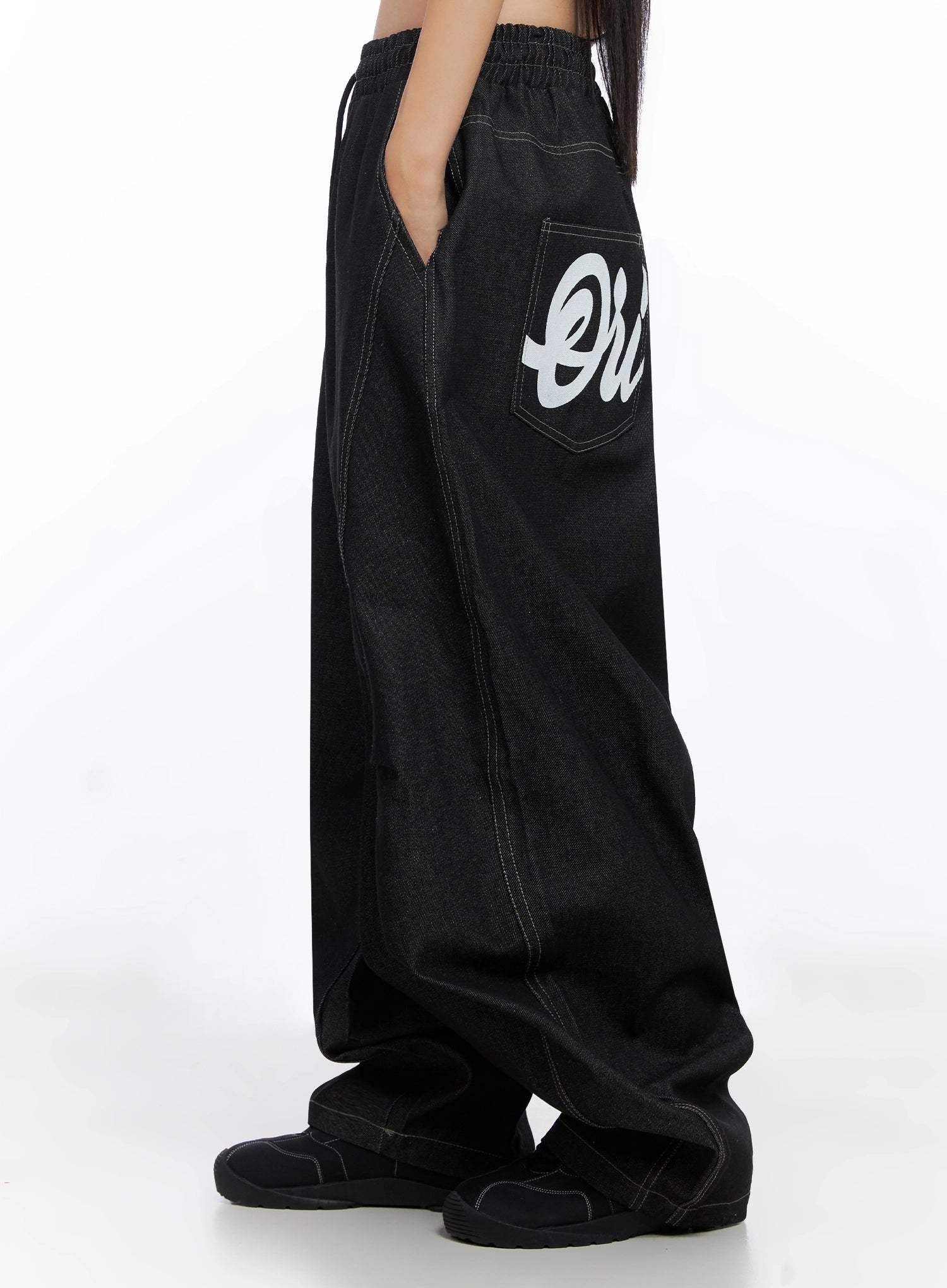 Ayra Graphic Baggy Jeans IO516 in Black, side close-up on model, curved wide-leg cotton denim, cursive lettering on back pocket, elastic waistband with drawstring, contrast white topstitching.