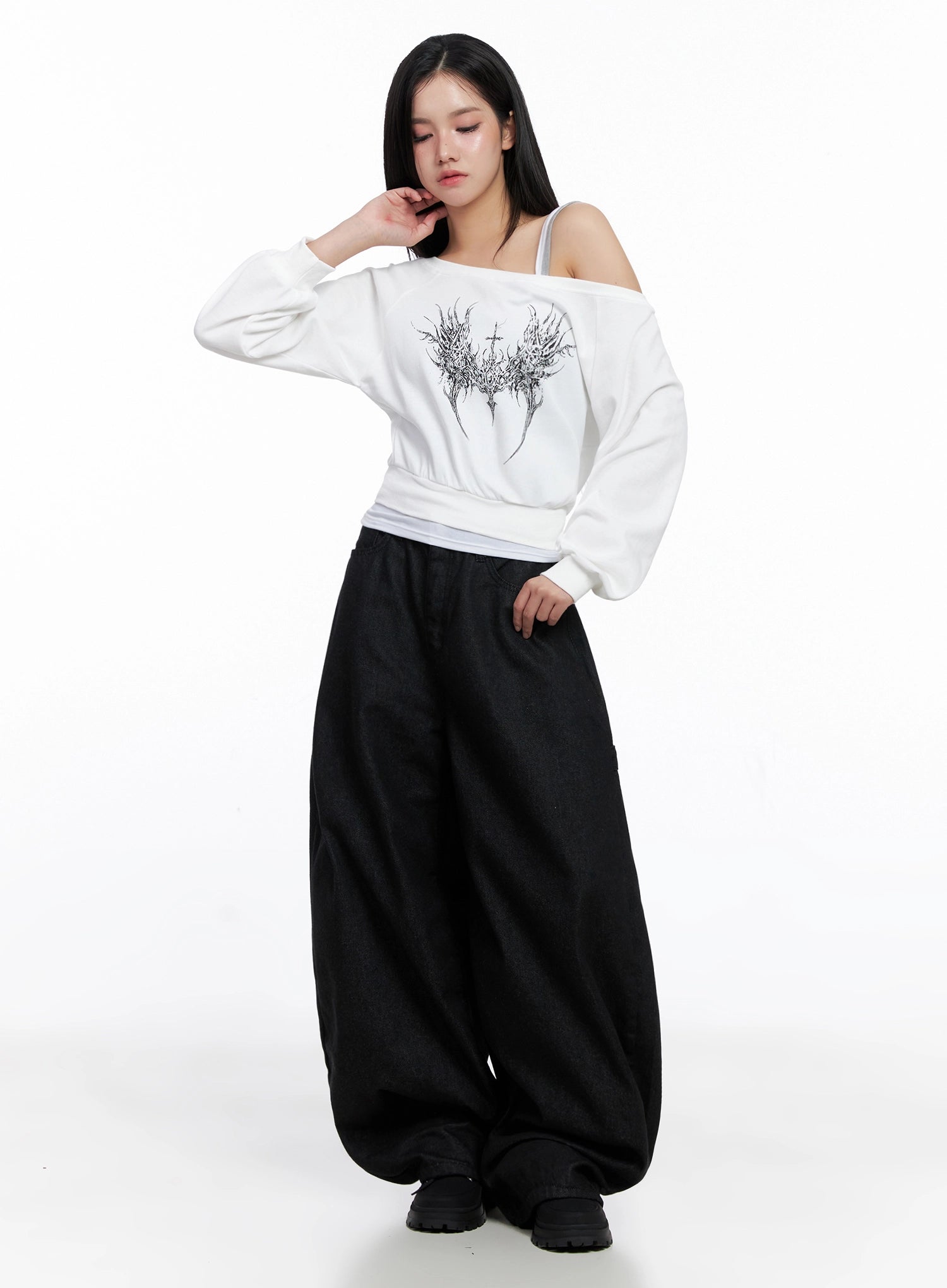 Lea Cross-Patch Baggy Jeans IS525 in black, front full-body editorial shot on model looking down, voluminous baggy cotton denim paired with white off-shoulder graphic sweatshirt, street acubi styling.