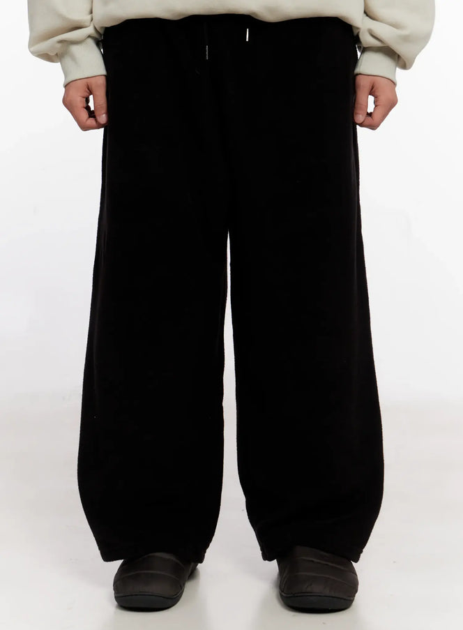 LEWKIN Men's Wide Leg Fleece Pants IO528 in black, front close-up on model, soft fleece fabric with relaxed wide-leg silhouette, drawstring waist, paired with cream sweatshirt and dark slides.