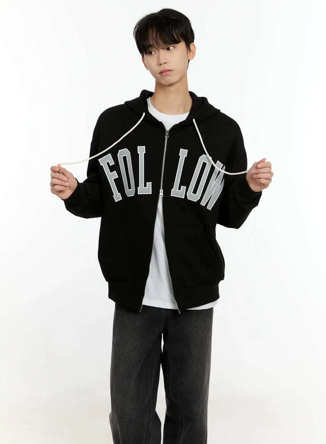 LEWKIN Men's Follow Me Zip-Up Hoodie IO531 in Black (M/L), front view on model holding drawstrings, oversized fit with arched FOLLOW appliqué lettering, layered over white tee with dark jeans.