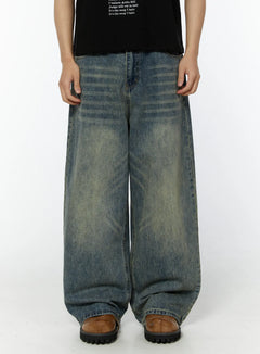 LEWKIN Men's Adan Baggy Washed Jeans IU508 in blue, front waist-to-hem crop on model, ultra-wide baggy leg with horizontal wash creases and vintage yellow-tint fading, paired with black tee and brown chunky sandals.