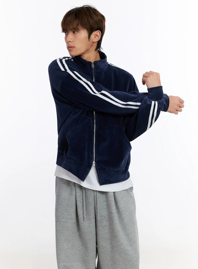 LEWKIN Men's Striped Track Jacket IN528 in dark blue, three-quarter side view on model, velour zip-up with white sleeve stripes, stand collar, arms crossed pose, layered over white tee with grey sweatpants.
