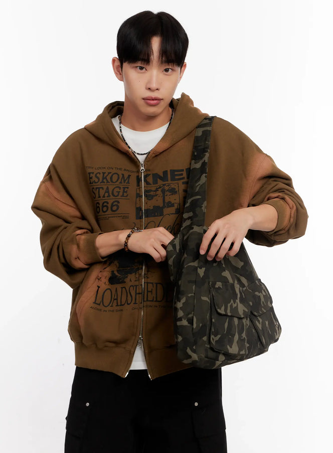 LEWKIN Camo Shoulder Bag with Utility Pockets IO 528 in dark green, front half-body shot on male model holding strap, camo canvas with flap pockets, styled with brown graphic zip-up hoodie and black cargo pants.