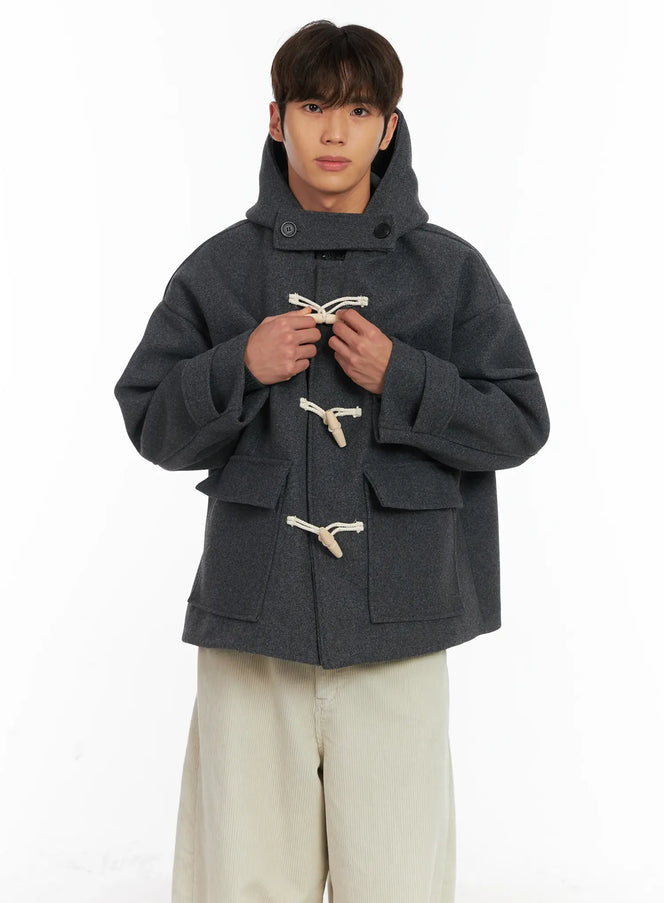 LEWKIN Men's Half Duffle Coat ID511 in charcoal grey, front close-up on model holding toggle closures, oversized wool-blend body with large hood and ivory rope toggles, paired with cream wide-leg trousers.
