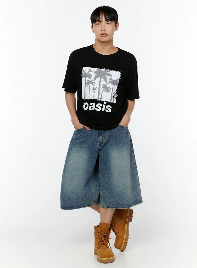 Men's Oasis Palm Graphic Tee IU515 in black, full-body front shot on model, relaxed fit short sleeve with oasis palm print, styled with wide denim bermuda shorts and tan boots.