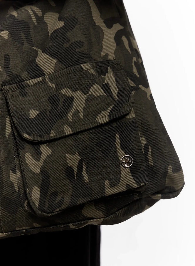 Camo Shoulder Bag with Utility Pockets IO 528 in dark green, macro close-up of front flap utility pocket, woven camo canvas texture and silver metal logo pin detail visible.