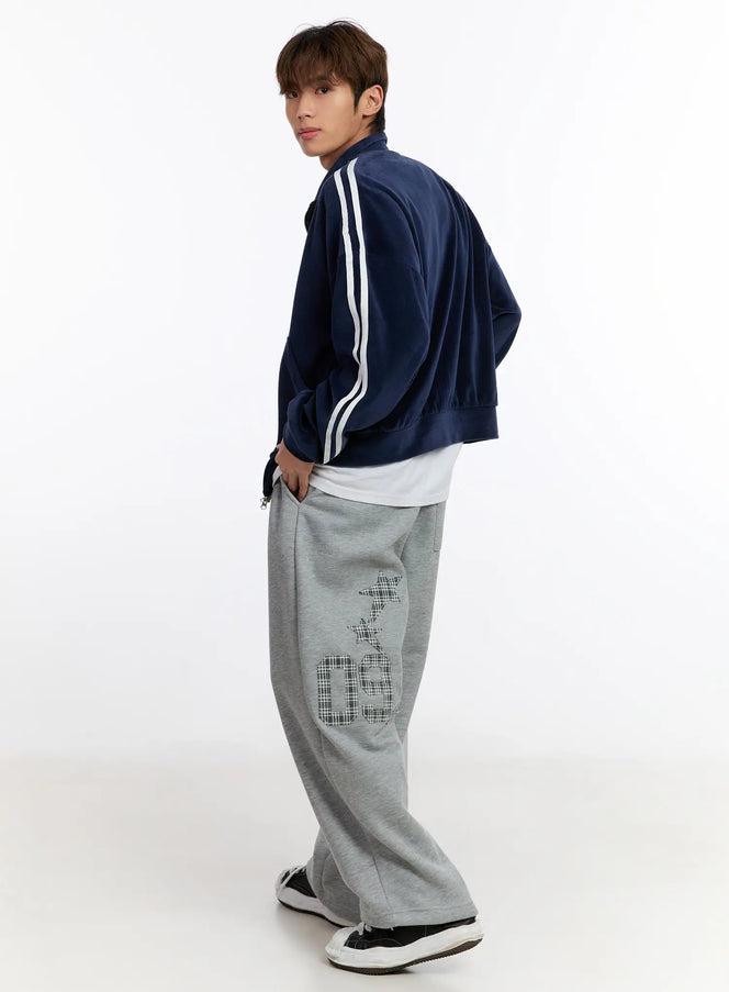 Men's Striped Track Jacket IN528 in dark blue, back three-quarter view on model glancing over shoulder, velour finish, white double sleeve stripes, layered over white tee with grey graphic sweatpants.