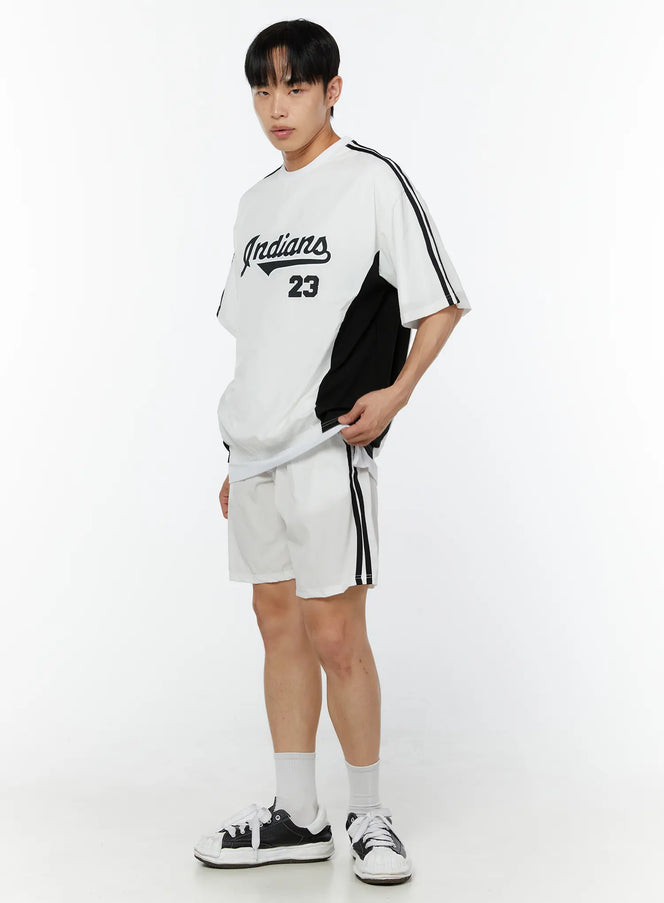 Men's Graphic Crewneck T-Shirt IU518 in white, three-quarter side view on model, oversized baseball tee with Indians 23 script graphic, black side panels and stripe sleeves, paired with white shorts and sneakers.
