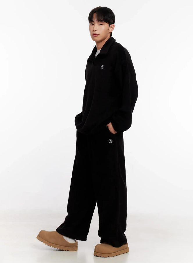 Men's Wide Leg Fleece Pants IO528 in black, three-quarter side view on model walking, full-length wide-leg fleece silhouette, LEWKIN logo embroidery, styled with matching black fleece top and tan boots.