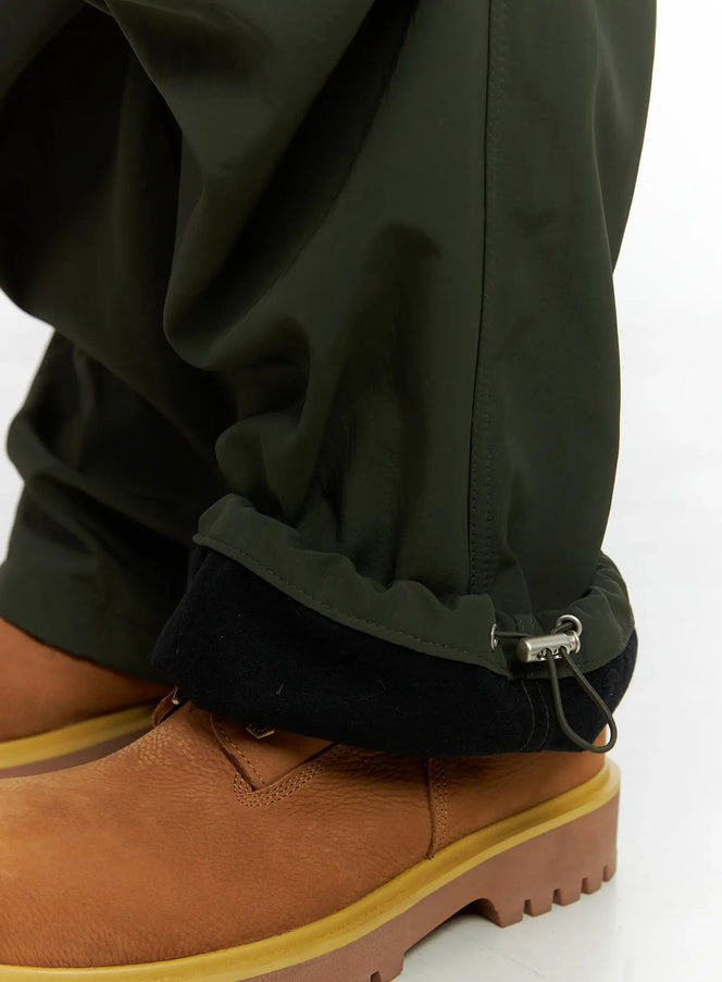 Men's Fleece Parachute Pants IN518 in dark green, hem detail close-up showing fleece-lined cuff and toggle drawstring adjuster, worn with tan boots.