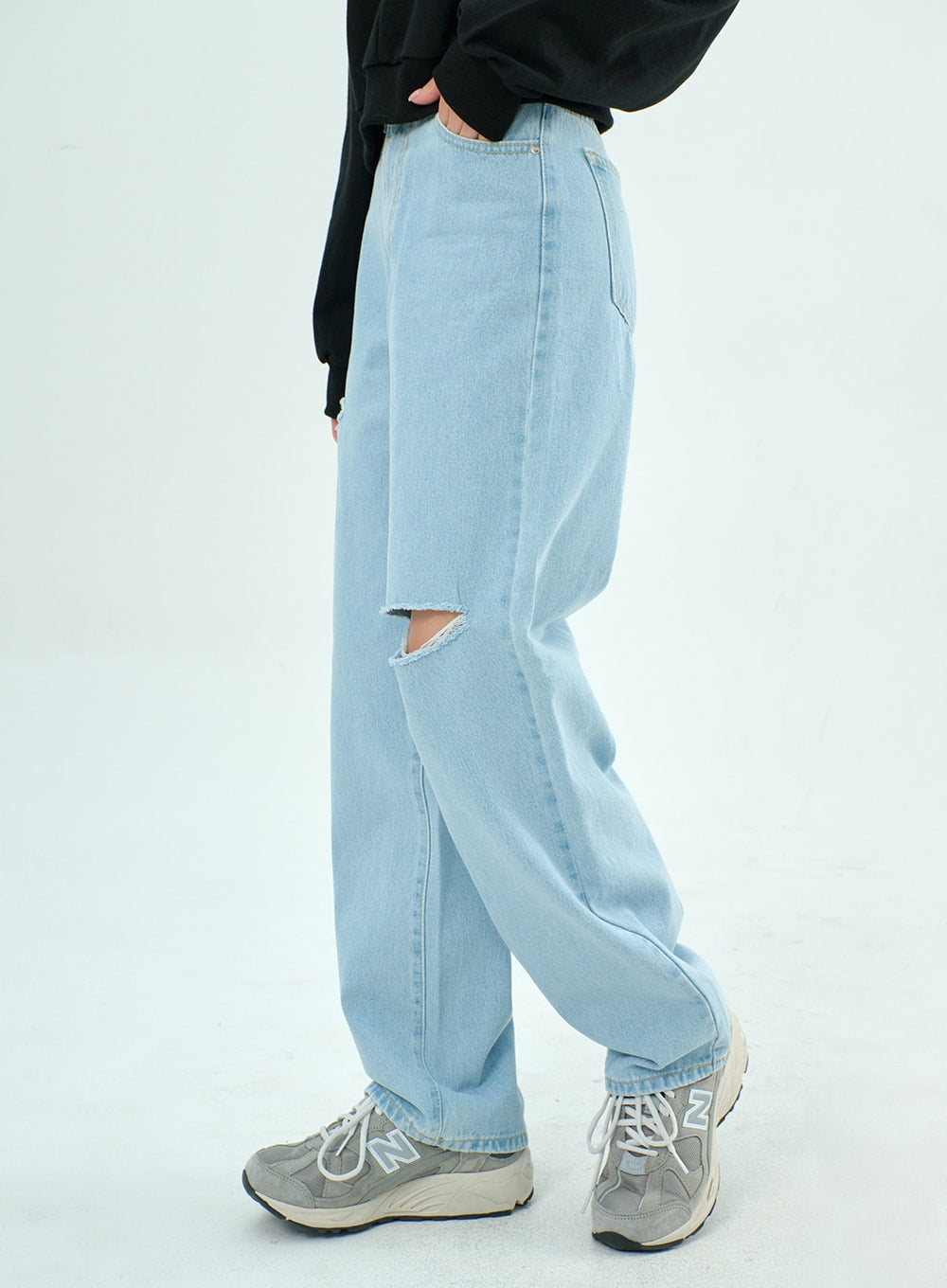 Ripped Basic Jeans BA328 in light blue, side view on model, wide-leg silhouette with open knee rip detail visible, styled with black sweatshirt and New Balance sneakers.