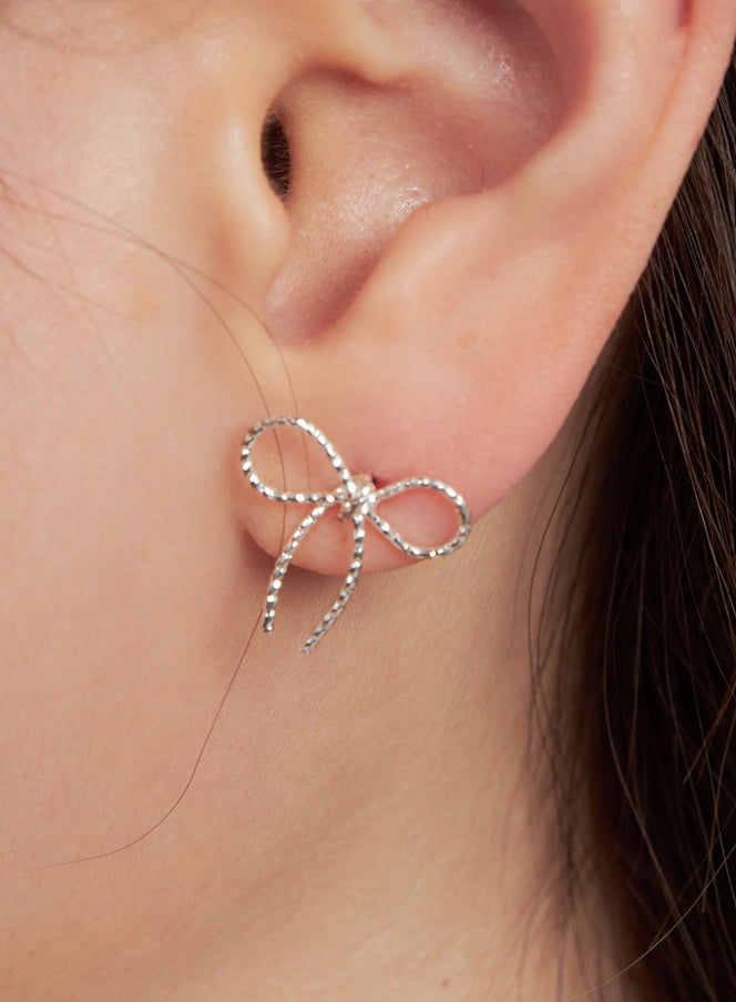 LEWKIN Noir Knot Ribbon Earrings IM531 in silver, extreme close-up worn on model's ear, open-wire bow silhouette with beaded texture and trailing tails.