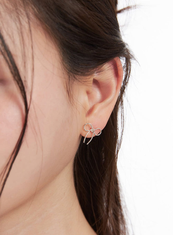 Noir Knot Ribbon Earrings IM531 in silver, side close-up on model, delicate open-wire ribbon bow stud with dangling tails against dark hair.