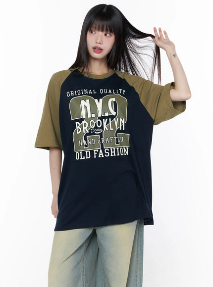 Casual Loose Fit Graphic Tee CU519 in dark blue, half-body front shot on model tossing hair, full Brooklyn NYC graphic print clearly visible, olive raglan short sleeves, crew neck, relaxed oversize fit.