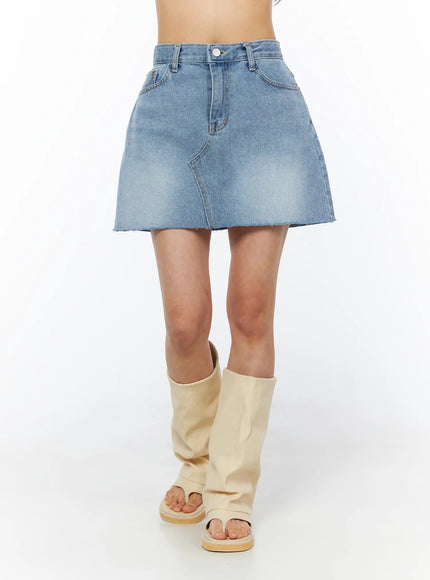 Casual Denim Mini Skirt CU522 in light blue, front close-up from waist to mid-thigh on model, asymmetric curved seam detail, button fly, faded wash, frayed raw hem, with cream boots.
