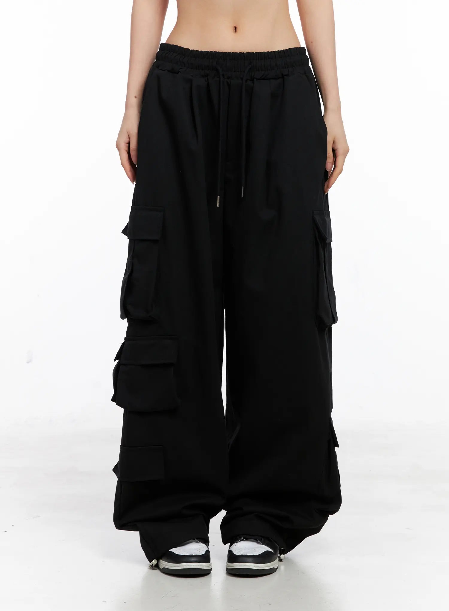 Wide Leg Cargo Pants CC1109 in black, front mid-shot on model, elastic drawstring waist, multiple flap cargo pockets stacked down both legs, long wide cotton fit, sneakers.