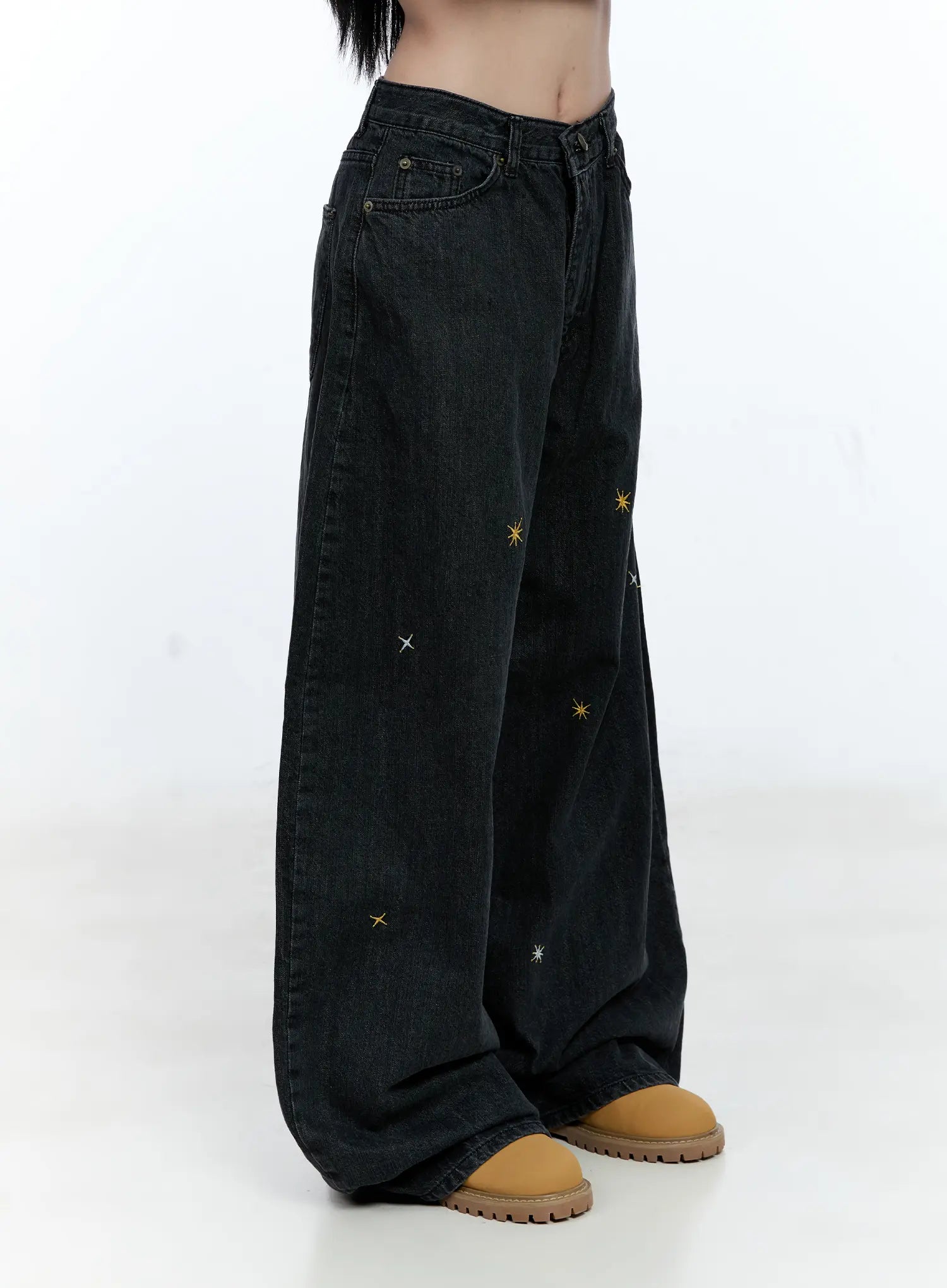 Wynter Star Embroidered Denim Jeans CG507 in black, side view cropped at waist, scattered gold and white star embroidery across wide-leg panels, mid-rise waistband detail, tan shoes.