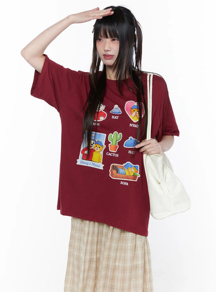 Oversized Graphic Print Tee CU519 in dark red, front upper-body view on model with hand raised, cartoon graphic print tee with round neck, paired with beige plaid skirt and white shoulder bag.