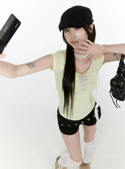 Angel Slim T-Shirt IL522 Light yellow front three-quarter view model posing with flip phone and bag from above slim ribbed scoop neck tee with glitter ANGEL CITY graphic