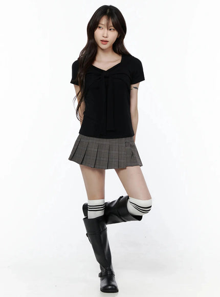 Bow Top IL506 Black front full-body shot on model with one leg raised, bow-tie chest detail, styled with gray pleated mini skirt and black knee-high boots