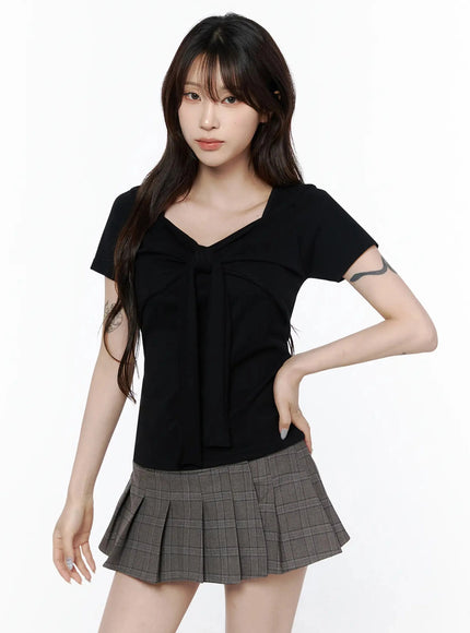 Bow Top IL506 Black front shot on model with hand on hip, relaxed pose showing bow-tie detail and fitted body, styled with gray pleated mini skirt