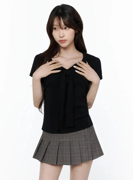 Bow Top IL506 Black front shot on model with both hands touching bow detail at chest, fitted short-sleeve top paired with gray pleated mini skirt