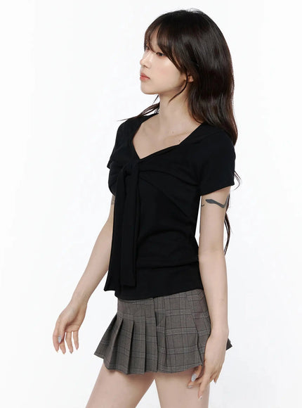 Bow Top IL506 Black side-front shot on model looking upward, showing bow-tie drape and slim waist, paired with gray pleated mini skirt