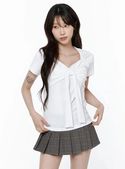 Bow Top IL506 White front shot on model with hands on waist, bow-tie chest detail, paired with gray pleated mini skirt