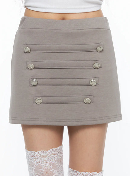 Buttoned Mini Skirt CL517 Beige close-up front detail shot on model highlighting four rows of silver military buttons and topstitched straps