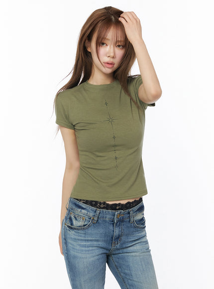 Chakra Graphic Slim Tee IL502 Dark green three-quarter front shot model raising hand to hair showing star graphic print slim cropped fit styled with jeans and black lace waistband