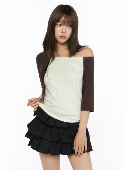 Color Block Off-Shoulder Tee IL523 Yellow front shot on model tugging hem, cream body with dark brown raglan half sleeves, off-shoulder neckline, paired with black tiered mini skirt