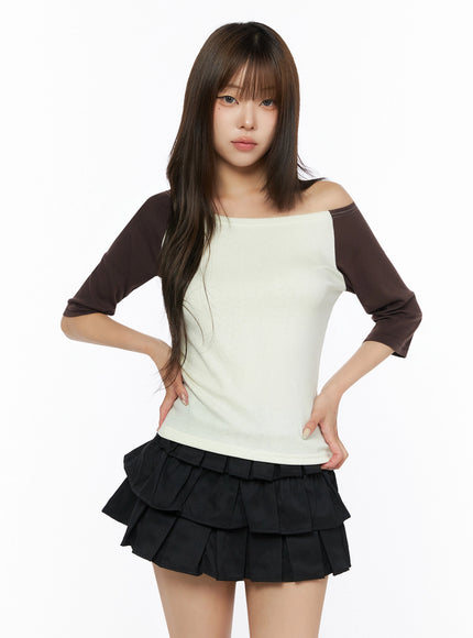 Color Block Off-Shoulder Tee IL523 Yellow front shot on model hands on hips, cream body with dark brown raglan sleeves, asymmetric off-shoulder neckline, contrast stitch detail visible