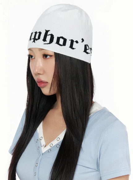 Contrast Graphic Text Reversible Beanie IL516 White three-quarter model shot, female in side-angled pose wearing gothic-lettered knit beanie with blue v-neck top