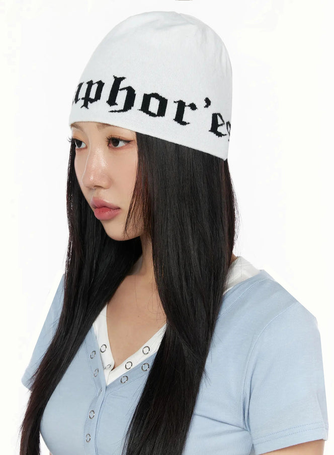 Contrast Graphic Text Reversible Beanie IL516 White three-quarter model shot, female in side-angled pose wearing gothic-lettered knit beanie with blue v-neck top