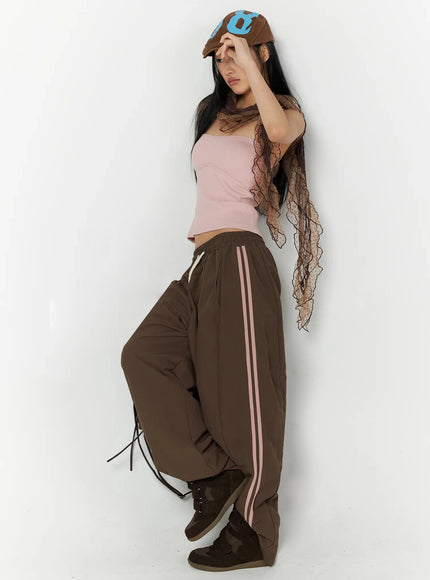 Contrast Stripe Track Pants IL509 Brown dynamic side-pose styled shot on model with cap and lace scarf, leg raised showing oversized wide-leg nylon silhouette