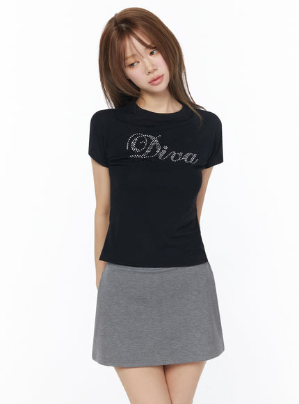 Diva Short Sleeve Tee IU525 Black front view on model with rhinestone script graphic, paired with gray mini skirt, slim fitted cotton tee