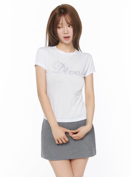 Diva Short Sleeve Tee IU525 White front view on model looking away, hands clasped at waist, rhinestone Diva graphic tee paired with gray mini skirt
